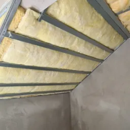 Niedokończona izolacja poddasza z wełny mineralnej w metalowej konstrukcji, surowe ściany przygotowane do dalszych prac wykończeniowych.