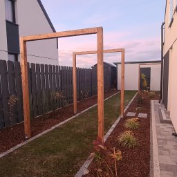 Złota rączka Mateusz Biały - Widok na ogród z drewnianą konstrukcją pergoli, ciemnoszarym ogrodzeniem panelowym i ścieżką z kostki brukowej, z widocznymi budynkami w tle.