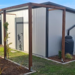 Złota rączka Mateusz Biały - Nowoczesny, blaszany domek narzędziowy z drewnianą pergolą, czarnym zbiornikiem na deszczówkę i ciemnym ogrodzeniem panelowym na tle zaoranego pola.