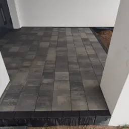 Ułożona kostka brukowa w odcieniach szarości przed wejściem do budynku, obramowana czarnym obrzeżem, widoczne białe filary wejścia i fragment niezagospodarowanego terenu.