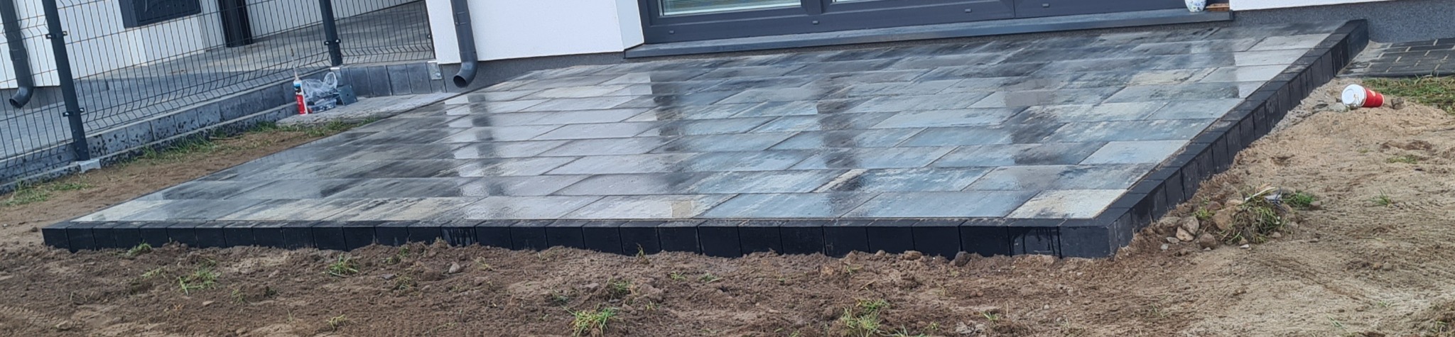 Nowo ułożona, wilgotna kostka brukowa w odcieniach szarości, obramowana czarnym obrzeżem, przylegająca do ściany budynku, z widocznym fragmentem trawnika i elementami krajobrazu w tle.