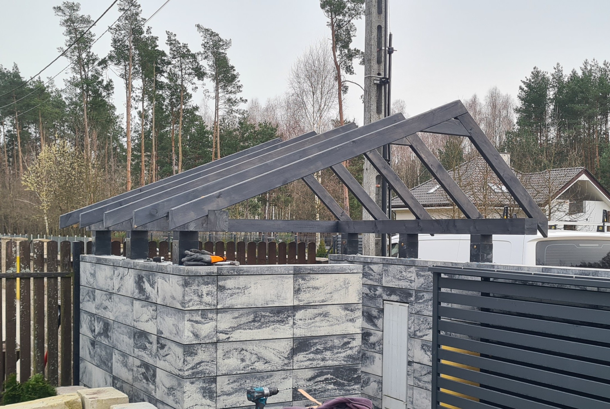 Konstrukcja z ciemnego drewna na murowanym słupku ogrodzenia z szarej kostki, widoczne narzędzia i rękawice robocze, w tle dom i drzewa.