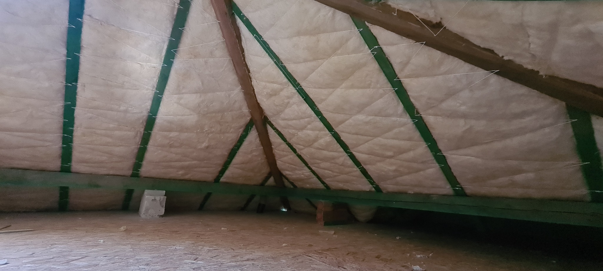 Ocieplenie poddasza wełną mineralną, widoczne zielone krokwie i białe sznurki mocujące izolację, surowa podłoga strychu.