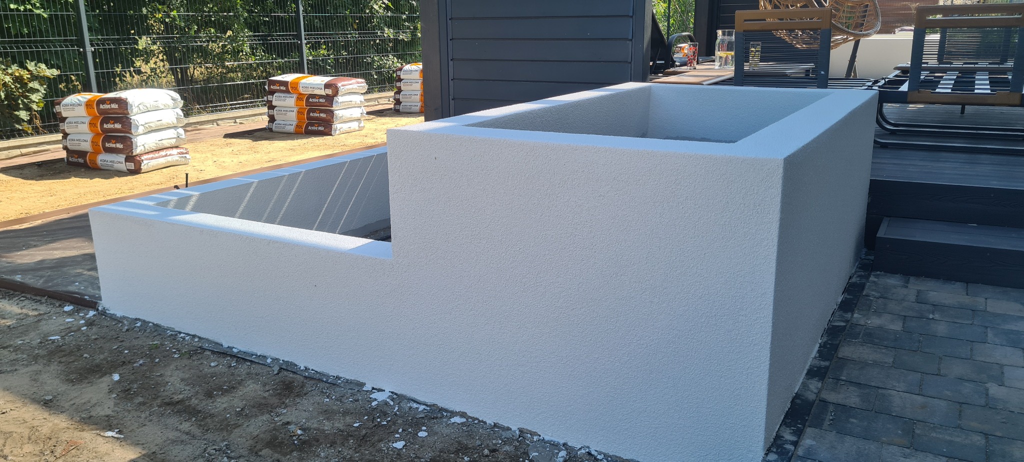 Nowoczesna, biała donica betonowa o dwóch poziomach, z workami ziemi w tle, umieszczona obok tarasu z desek kompozytowych i kostki brukowej.