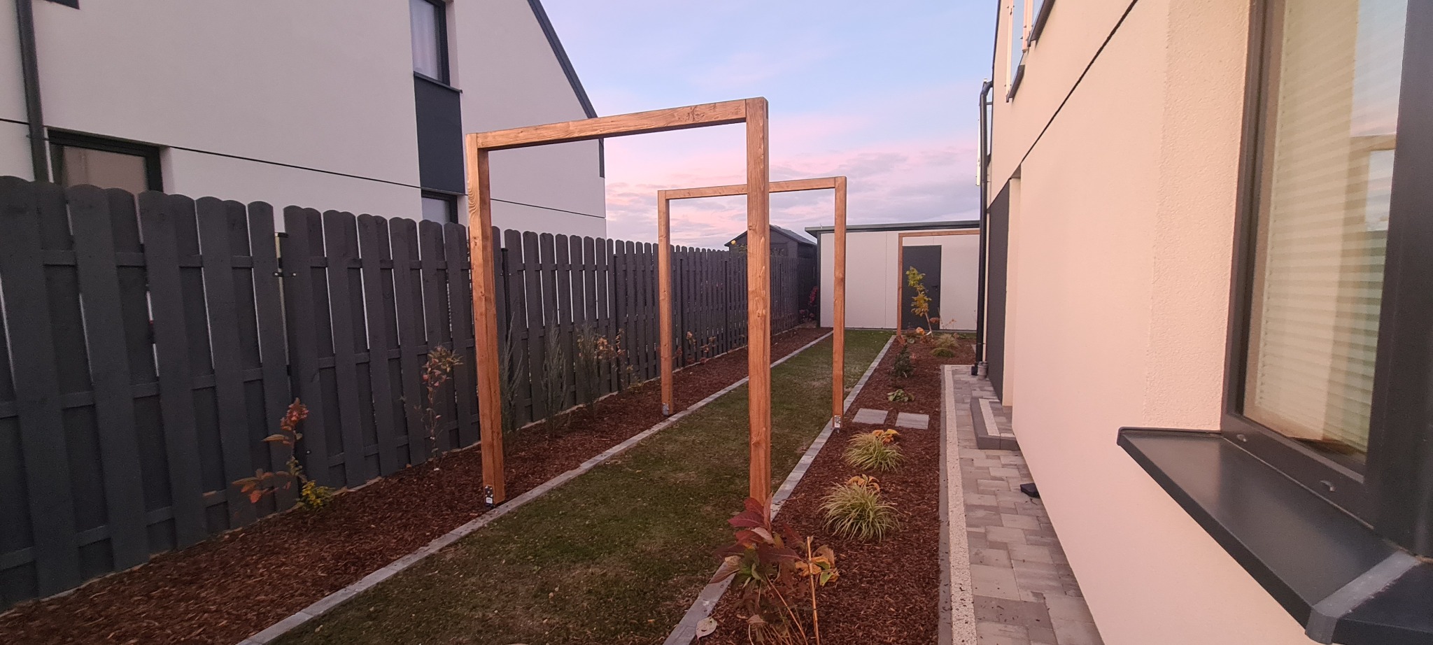 Widok na ogród z drewnianą konstrukcją pergoli, ciemnoszarym ogrodzeniem panelowym i ścieżką z kostki brukowej, z widocznymi budynkami w tle.