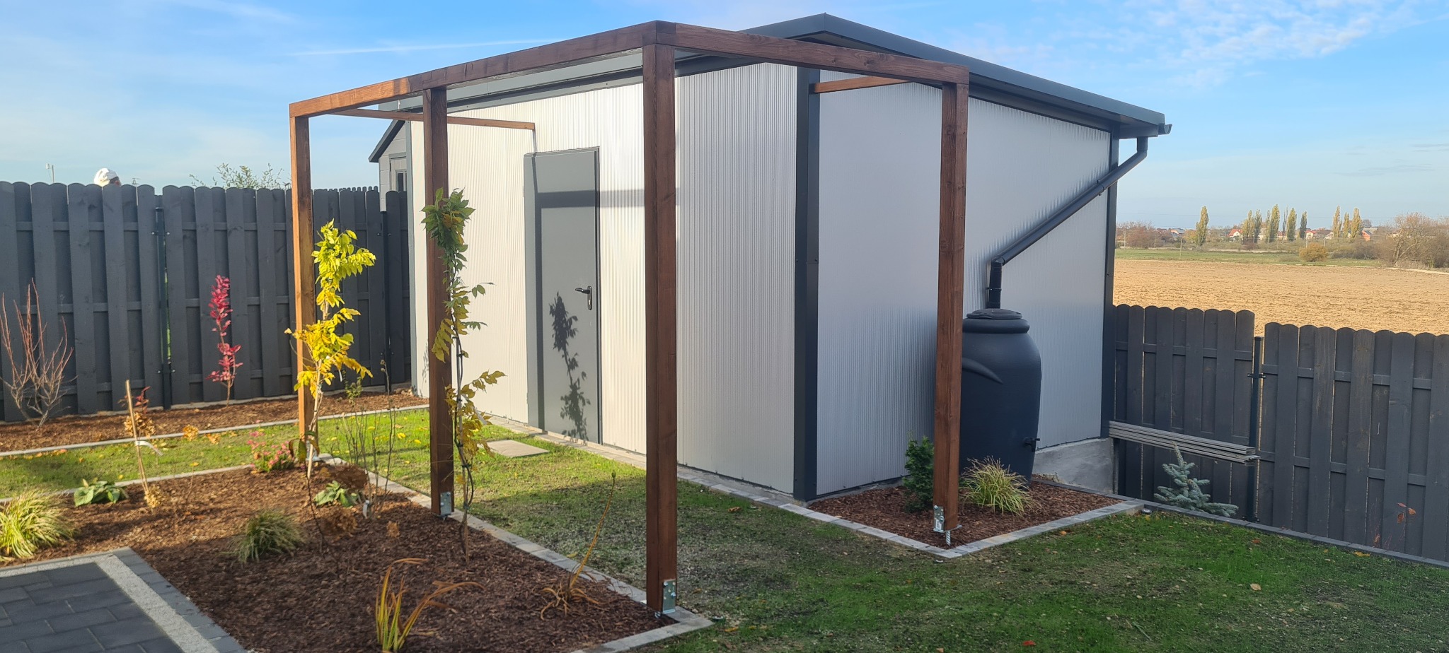 Nowoczesny, blaszany domek narzędziowy z drewnianą pergolą, czarnym zbiornikiem na deszczówkę i ciemnym ogrodzeniem panelowym na tle zaoranego pola.