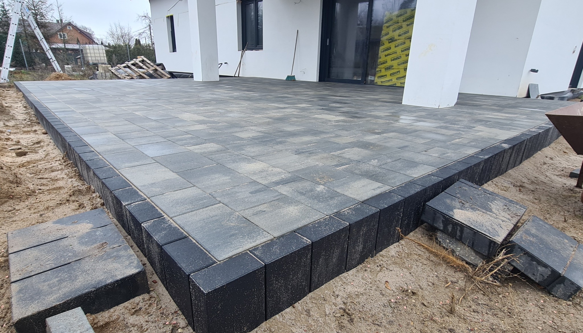 Nowo ułożony taras z szarej kostki brukowej z czarnym obrzeżem, widoczne pozostałości piasku i zaplecze budowy.