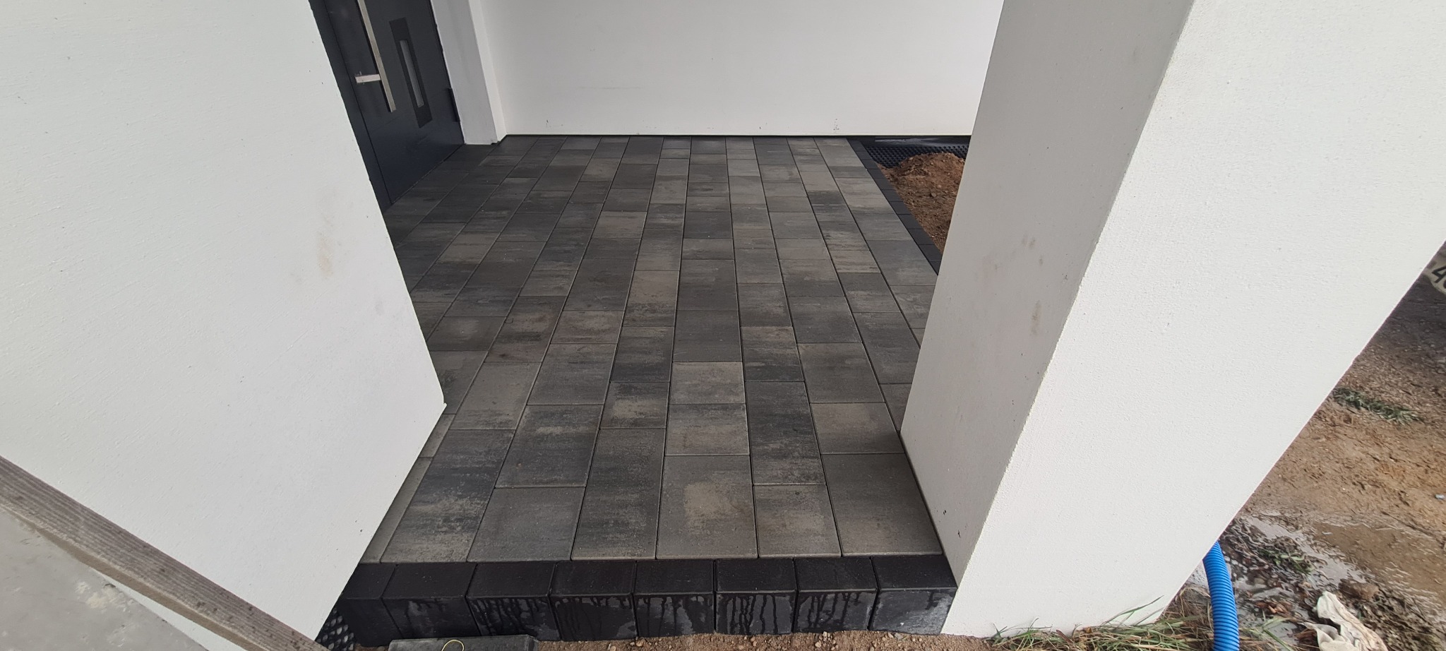 Ułożona kostka brukowa w odcieniach szarości przed wejściem do budynku, obramowana czarnym obrzeżem, widoczne białe filary wejścia i fragment niezagospodarowanego terenu.