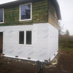 PWMS-BUD SPÓŁKA Z OGRANICZONĄ ODPOWIEDZIALNOŚCIĄ - Budynek w trakcie prac przygotowawczych do ocieplenia elewacji, widoczne drewniane deski pokryte folią ochronną, okna w białych ramach, wstępny etap prac.