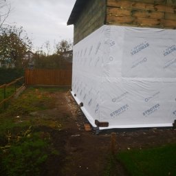 PWMS-BUD SPÓŁKA Z OGRANICZONĄ ODPOWIEDZIALNOŚCIĄ - Budynek w trakcie ocieplania, zabezpieczony białą folią paroizolacyjną z nadrukowanym logo producenta, widoczne drewniane elementy konstrukcji oraz okno z białym obramowaniem, w tle ogrodzenie...