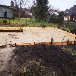 PWMS-BUD SPÓŁKA Z OGRANICZONĄ ODPOWIEDZIALNOŚCIĄ - Przygotowanie terenu pod wylewkę fundamentów: drewniany szalunek wypełniony piaskiem, widoczna pryzma ciemnej ziemi, w tle zabudowa jednorodzinna.