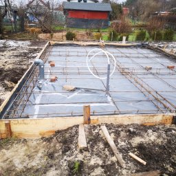 PWMS-BUD SPÓŁKA Z OGRANICZONĄ ODPOWIEDZIALNOŚCIĄ - Zbrojenie fundamentów pod budowę domu, szalunek z desek, folia izolacyjna, rury kanalizacyjne wystające ponad poziom gruntu, w tle budynek gospodarczy.