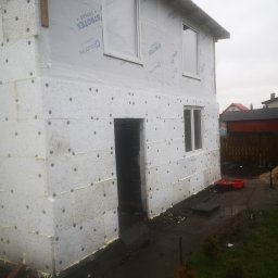 PWMS-BUD SPÓŁKA Z OGRANICZONĄ ODPOWIEDZIALNOŚCIĄ - Surowy stan budynku w trakcie ocieplania elewacji styropianem z widoczną folią wiatroizolacyjną marki Strotex, świeżo wstawione okna, ziemia wokół budynku.