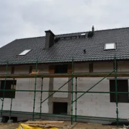 Dom w trakcie budowy z białych bloczków, z czarnym dachem i kominem, otoczony rusztowaniami. Widoczne okna dachowe i otwory okienne.