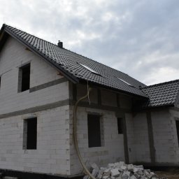 PWMS-BUD SPÓŁKA Z OGRANICZONĄ ODPOWIEDZIALNOŚCIĄ - Dwukondygnacyjny dom w stanie surowym otwartym z czarną dachówką, widoczne pustaki, nadproża, fragment elewacji i składowane materiały budowlane.