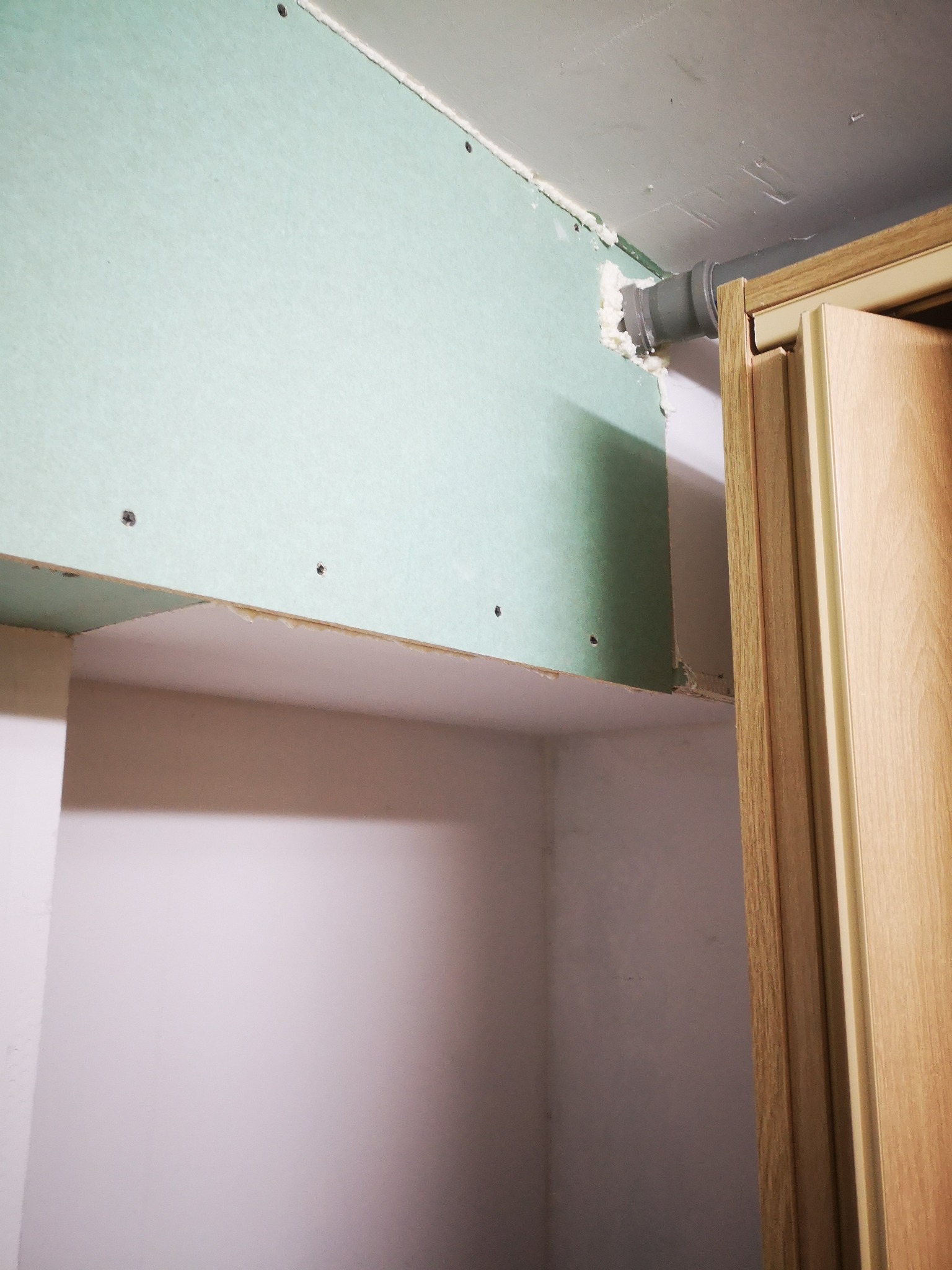 Fragment ściany z zieloną płytą gipsowo-kartonową przymocowaną do sufitu, widoczna szpara wypełniona pianką montażową, szara rura wystająca ze ściany obok drewnianej futryny drzwi.