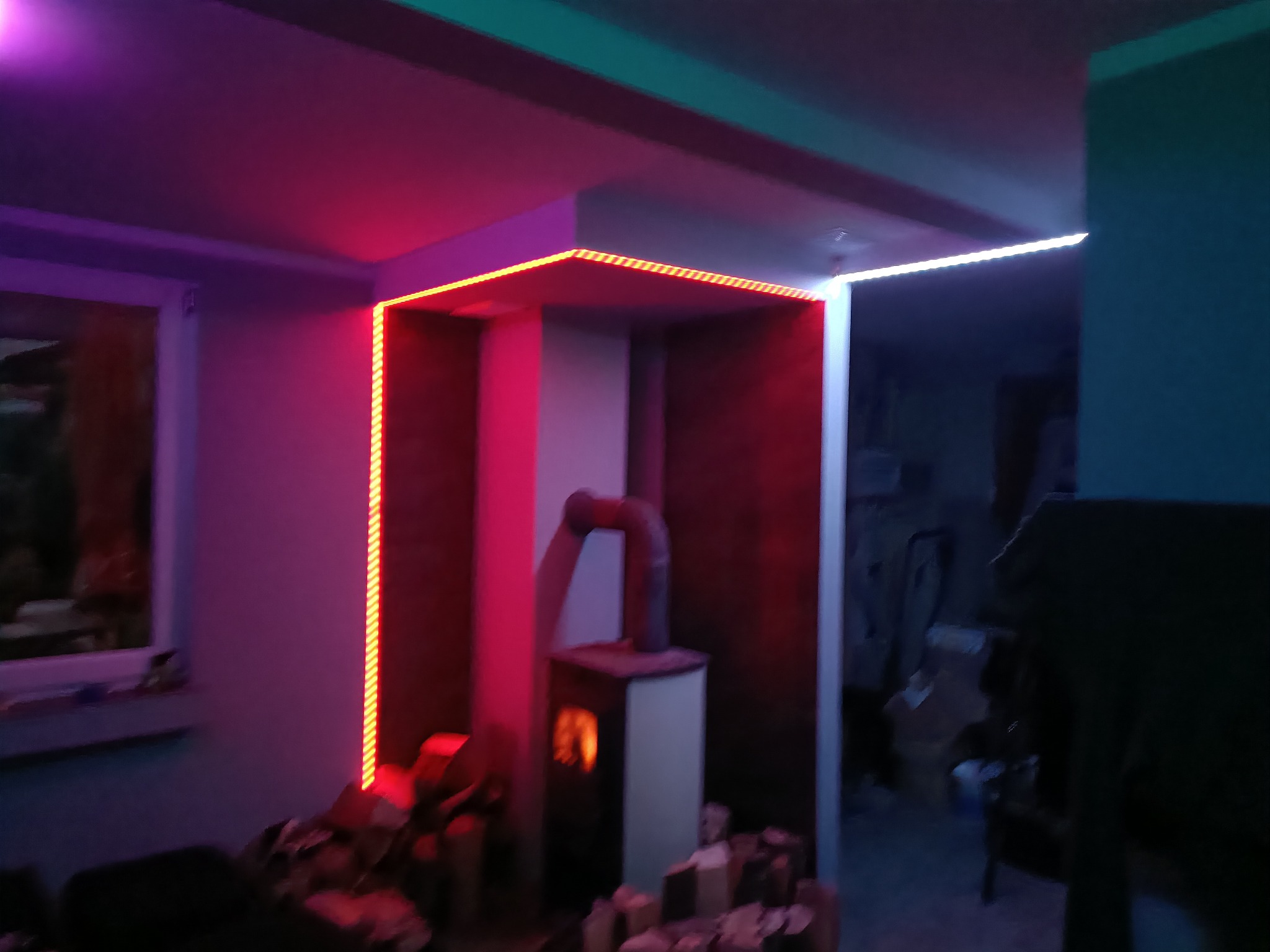 Pokój z kominkiem i dekoracyjnym oświetleniem LED w kolorach różowym i turkusowym, z widocznym fragmentem okna po lewej stronie.