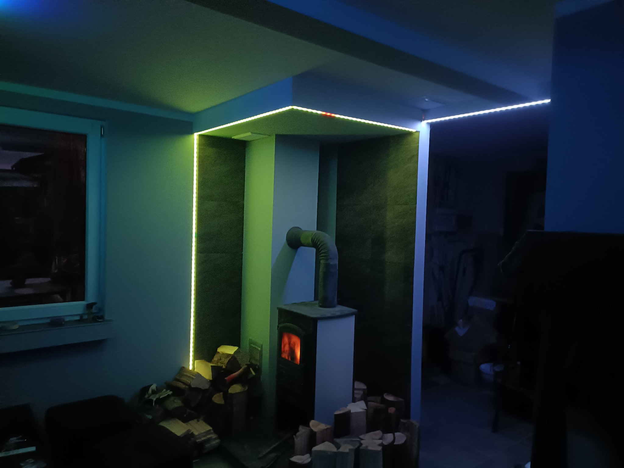 Wnętrze pokoju z kominkiem, oświetlone ledowymi taśmami w kolorach żółtym i niebieskim, z ułożonym drewnem obok.