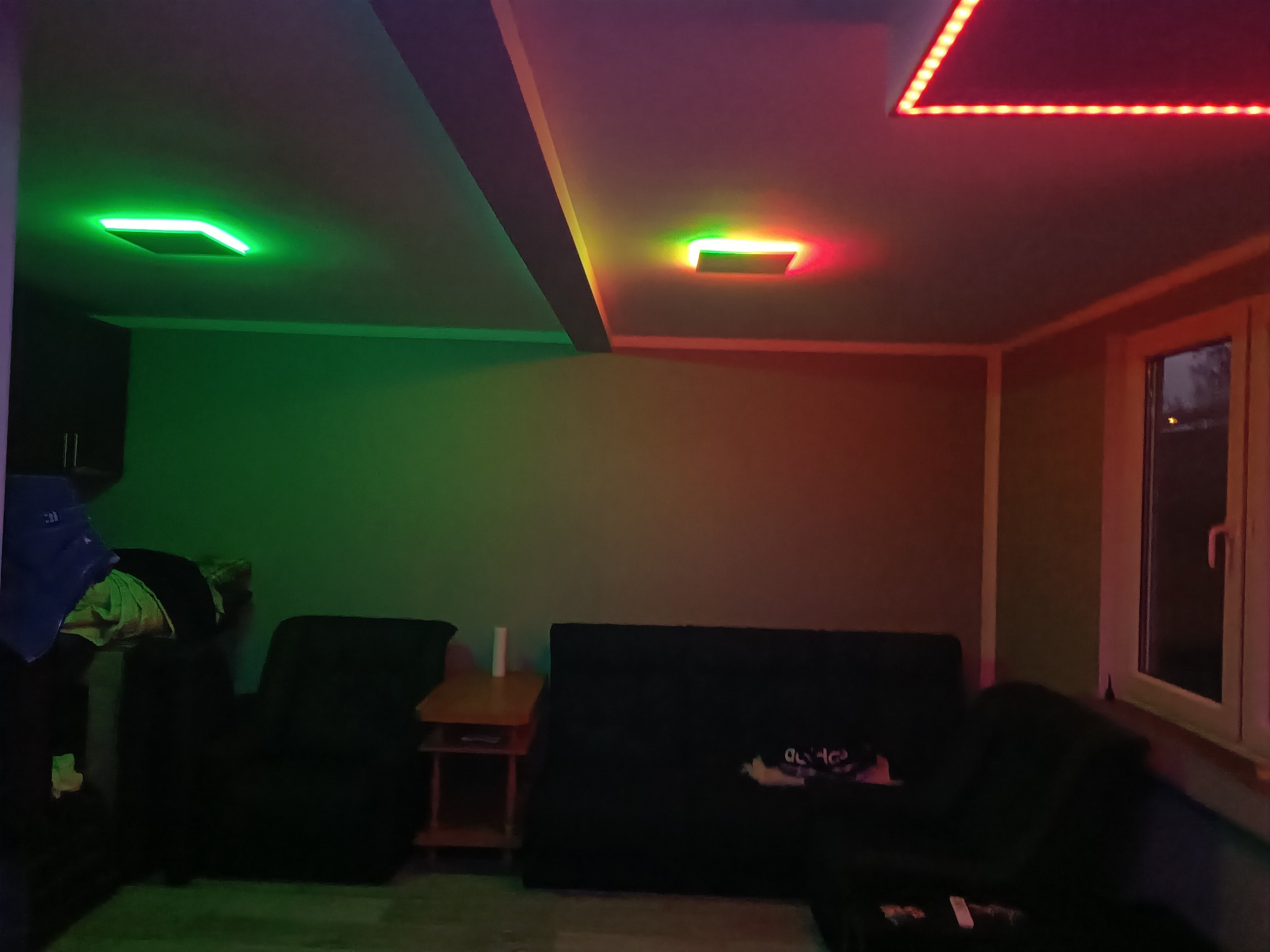 Pokój z sufitem podwieszanym z oświetleniem LED: zielone panele w lewej części i czerwone linie po prawej, z ciemnymi meblami w tle.