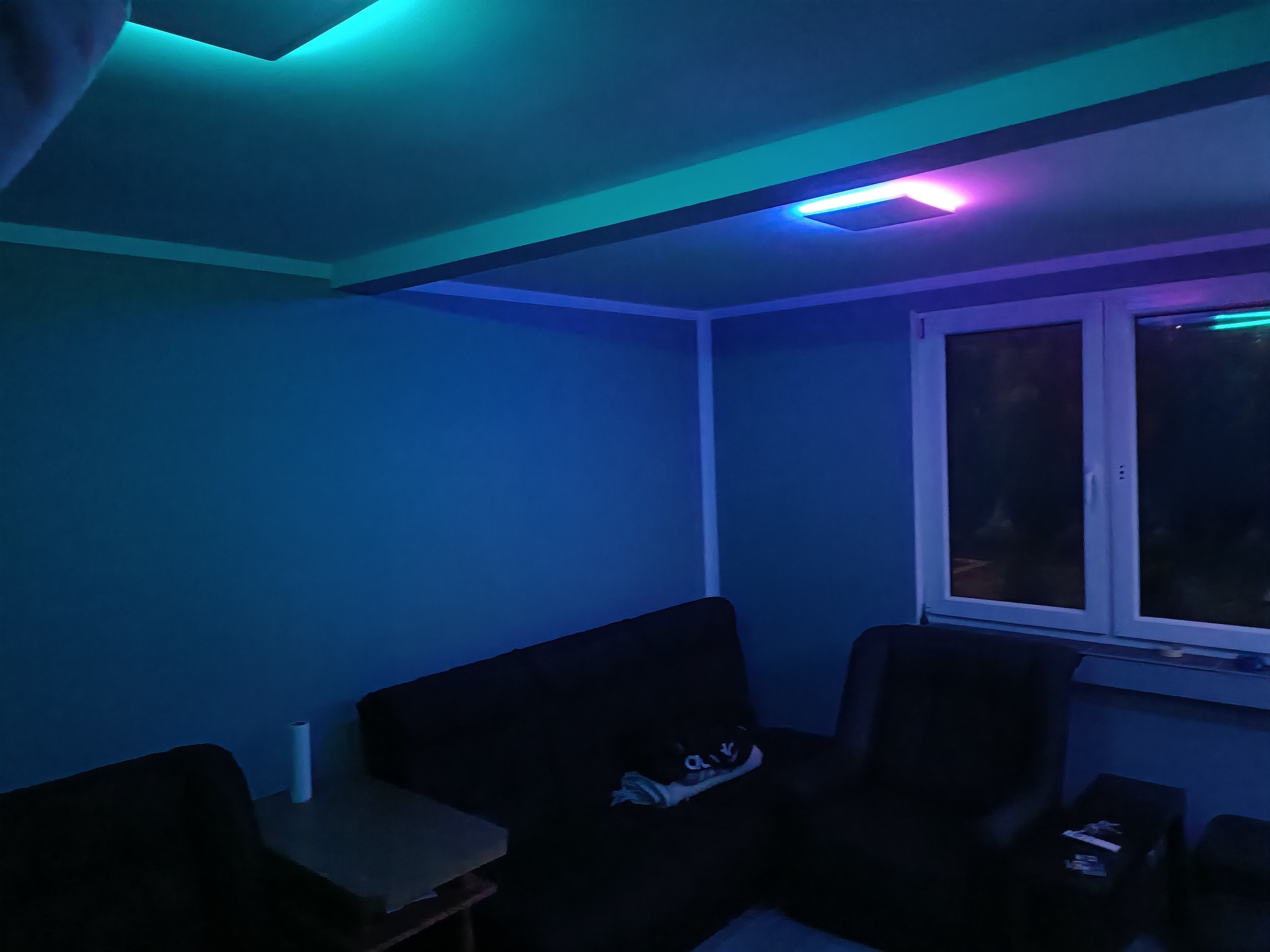 Pokój z oświetleniem LED RGB: turkusowe i różowe światło na suficie, szare ściany, ciemne meble wypoczynkowe, widok na okno nocą.