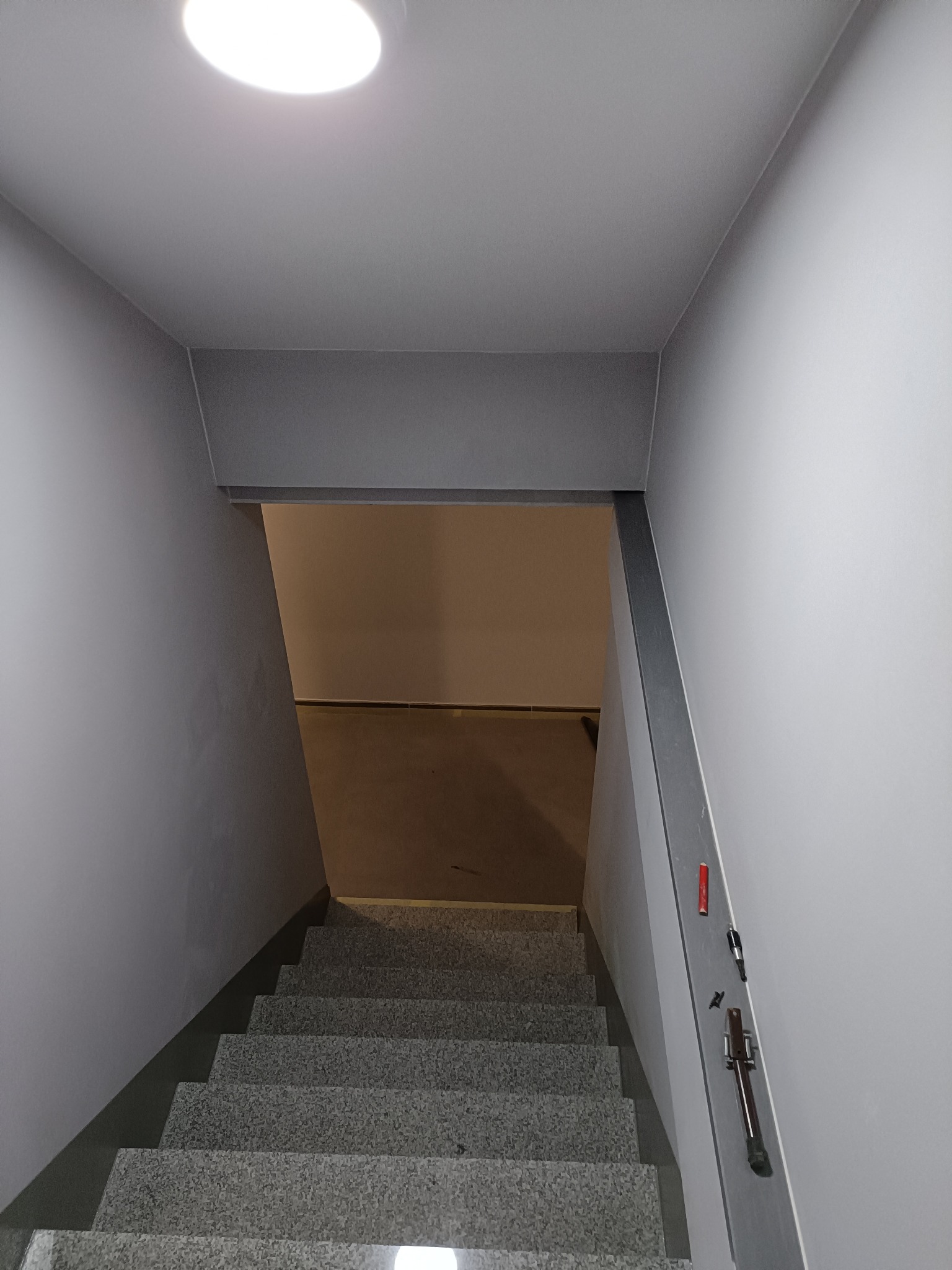 Schody ze ścianami pomalowanymi na szaro, widok z góry, z widocznym oświetleniem sufitowym i narzędziami przymocowanymi do ściany.