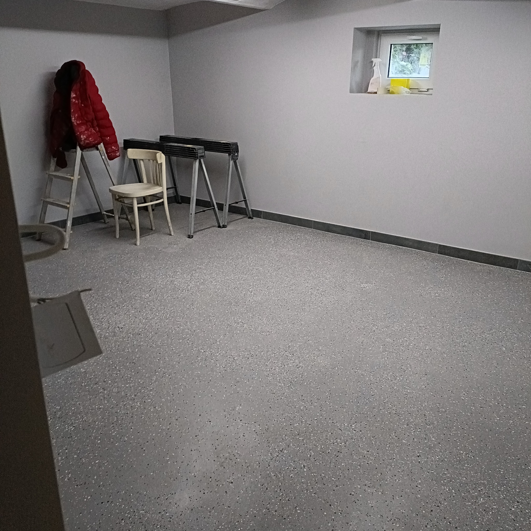 Szare pomieszczenie po remoncie z nową, szarą podłogą z drobnymi plamkami, drabina, krzesło i kozły malarskie pod ścianą, okno z widocznymi środkami czystości.