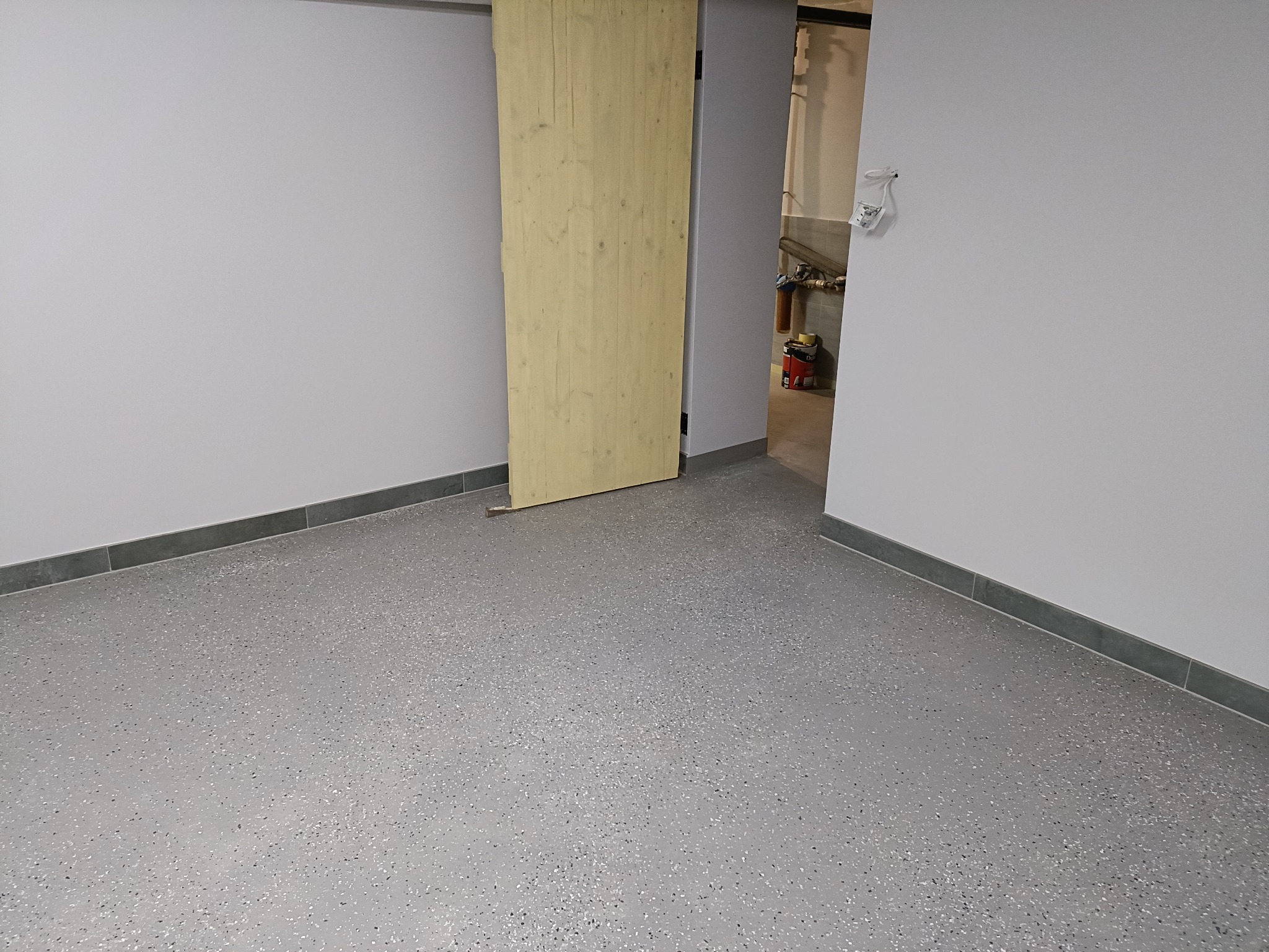 Szara podłoga żywiczna z drobnymi kolorowymi wtrąceniami, drewniane przesuwne drzwi do pomieszczenia z instalacjami, szare ściany z listwami przypodłogowymi.