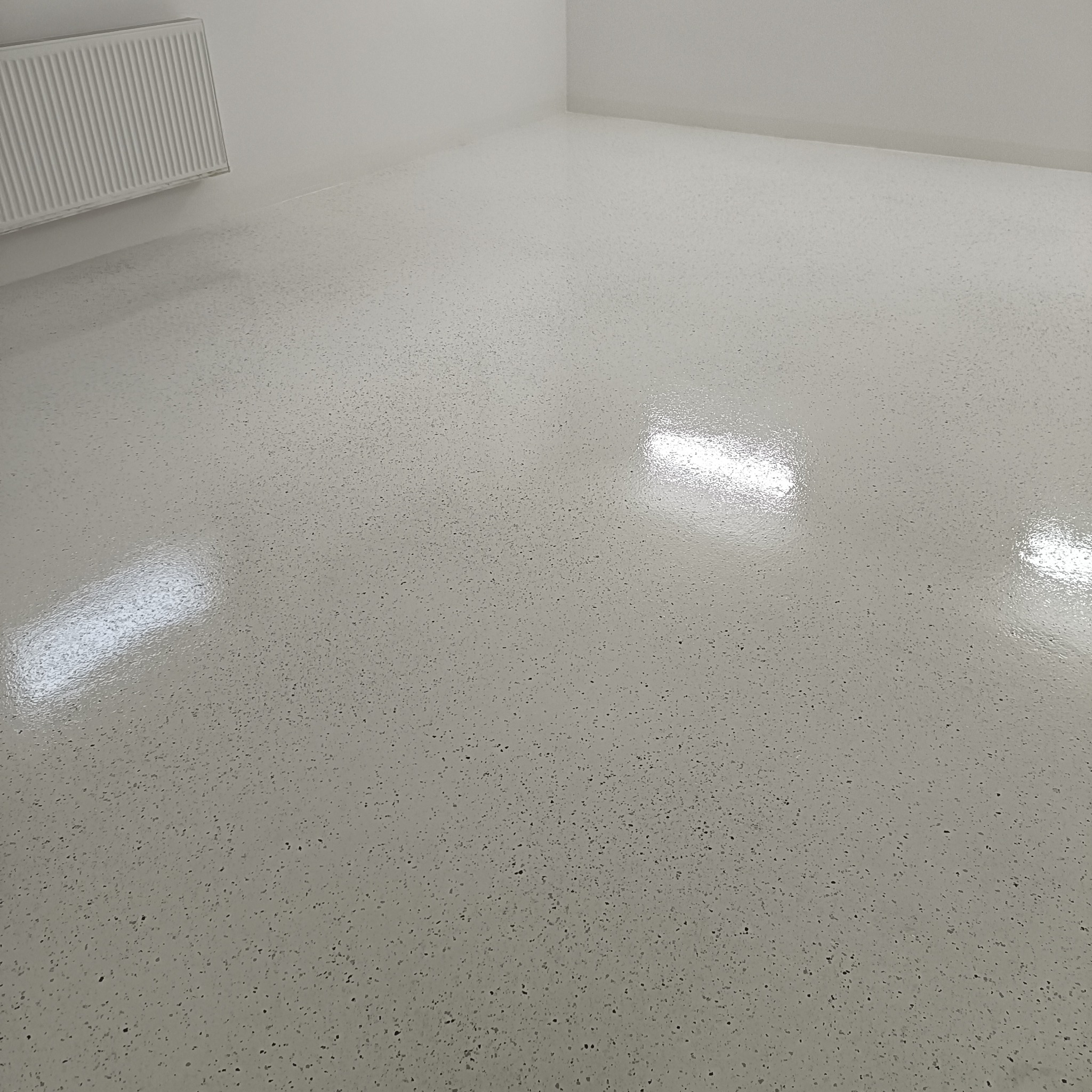 Jasnoszara, błyszcząca posadzka żywiczna z drobnymi, ciemnymi inkluzjami w pustym, białym pomieszczeniu z grzejnikiem na ścianie.