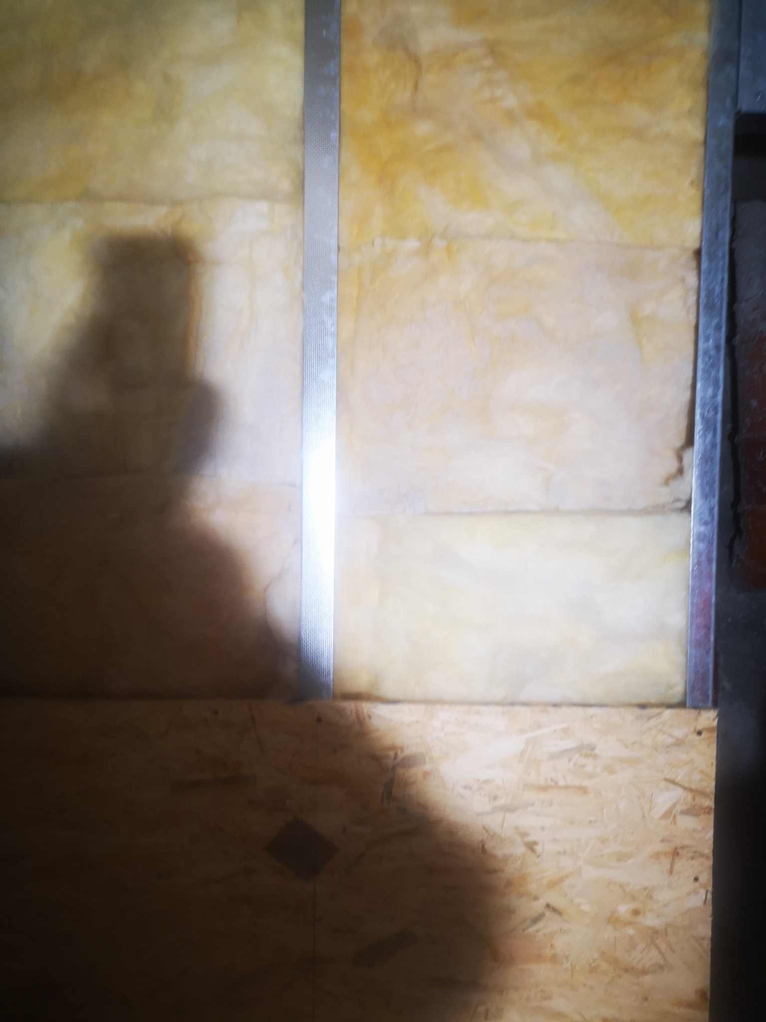 Ściana z izolacją z wełny mineralnej, częściowo zakryta płytą OSB, widoczny metalowy profil i cień osoby.