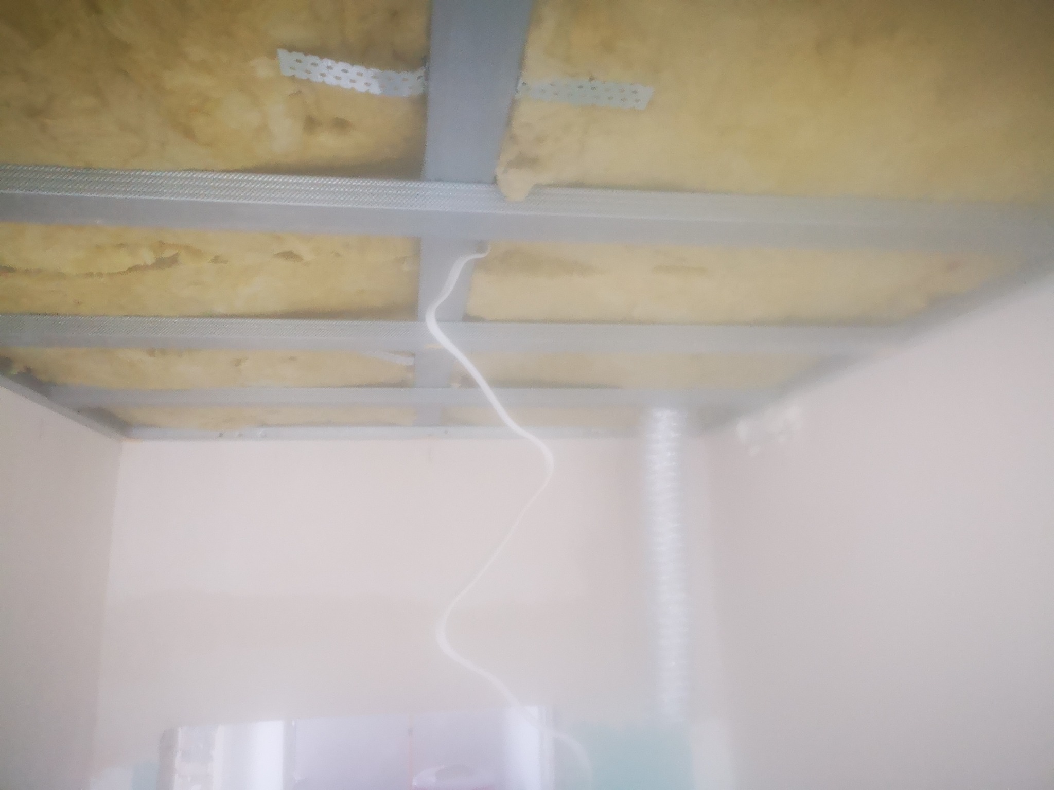 Sufit podwieszany w trakcie montażu: metalowa konstrukcja z profilem CD i UD, ocieplenie z wełny mineralnej, wystający przewód elektryczny, widok z dołu.