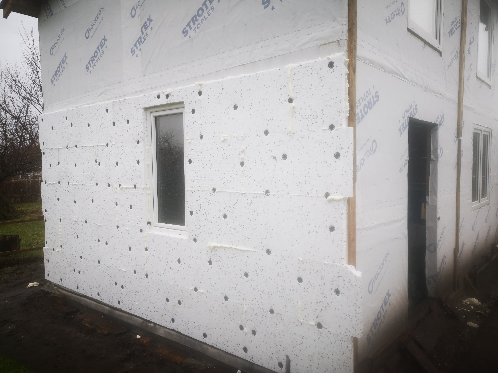 Ściana domu w trakcie ocieplania styropianem z widocznymi kołkami montażowymi i pianką uszczelniającą, zabezpieczona folią wiatroizolacyjną z nadrukowanym logo producenta, widok z dołu.