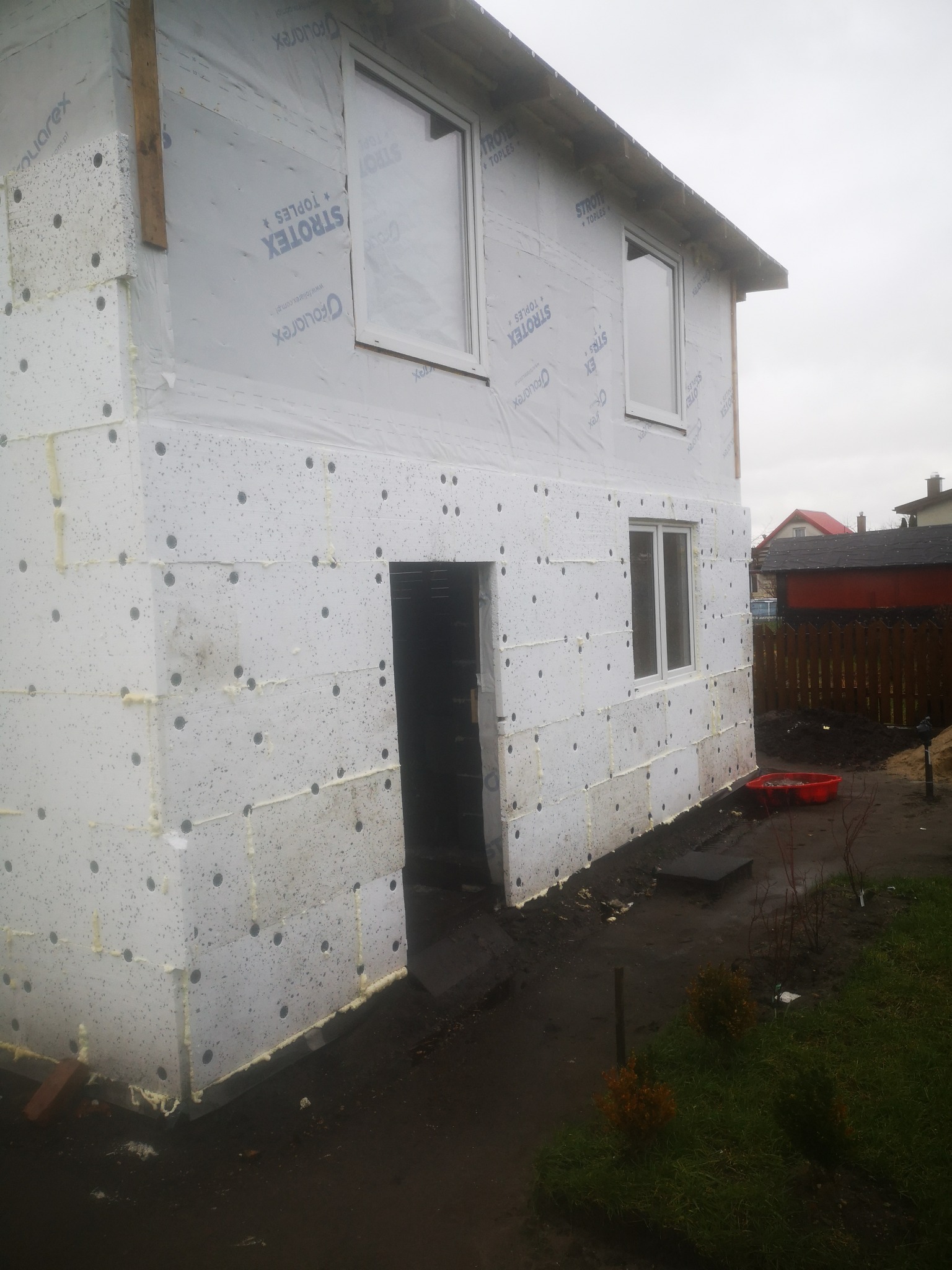 Surowy stan budynku w trakcie ocieplania elewacji styropianem z widoczną folią wiatroizolacyjną marki Strotex, świeżo wstawione okna, ziemia wokół budynku.