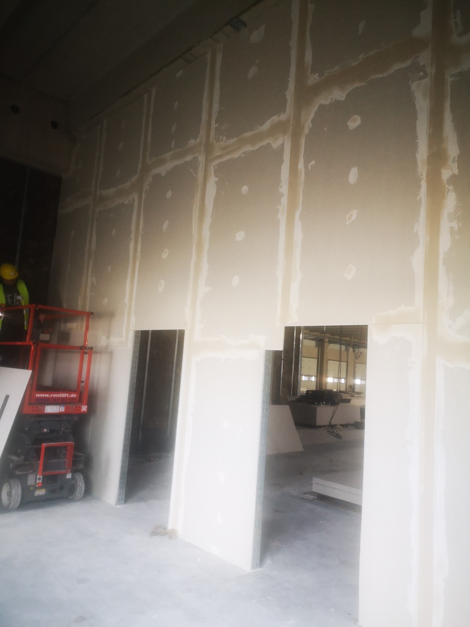 Montaż płyt karton-gips na ścianie z widocznymi spoinami, pracownik na podnośniku koszowym w żółtym kasku, surowy stan wykończenia, widok na sąsiednie pomieszczenie przez otwór w ścianie.