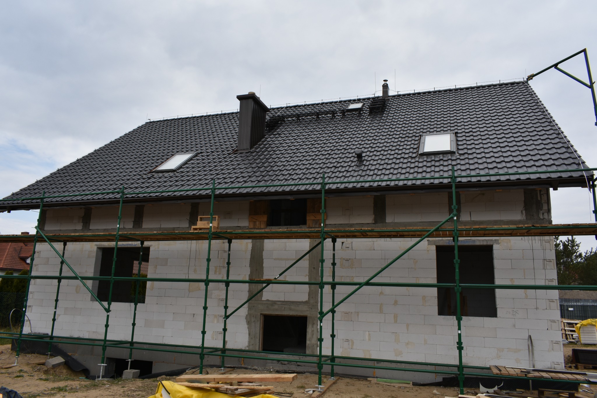 Dom w trakcie budowy z białych bloczków, z czarnym dachem i kominem, otoczony rusztowaniami. Widoczne okna dachowe i otwory okienne.
