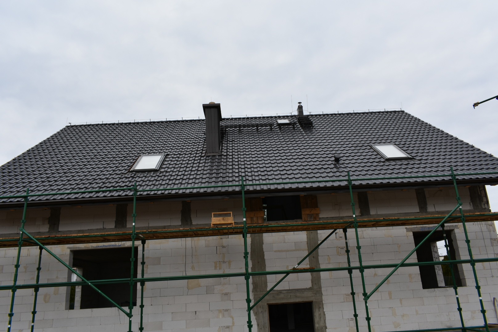 Dom w trakcie budowy: białe ściany z pustaków, czarny dach z lukarnami, zielone rusztowania. Widok na fragment budynku z góry, pochmurne niebo w tle.