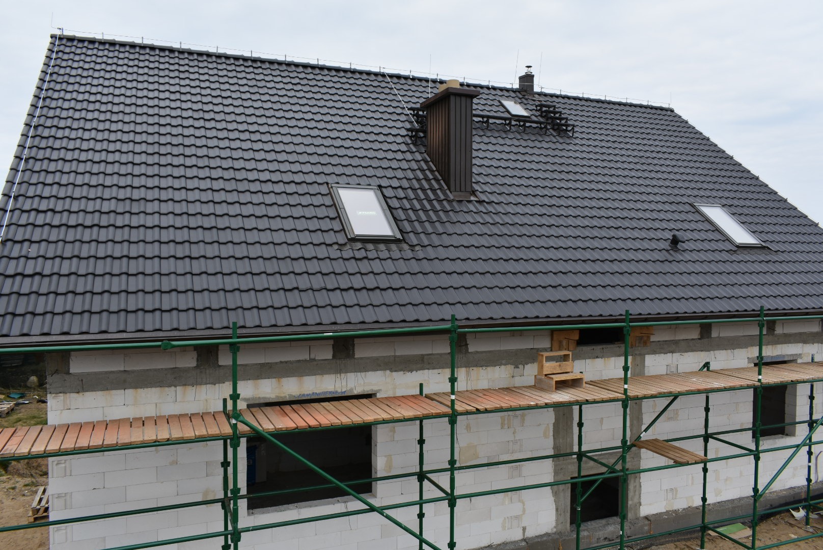 Dom w trakcie budowy z czarną dachówką, widoczne okna dachowe, komin i rusztowanie. Ściany z białego bloczka, częściowo ocieplone. Pochmurne niebo w tle.