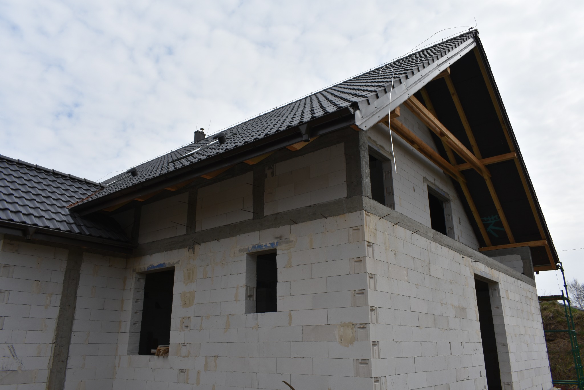 Surowy dom z białego betonu komórkowego, czarny dach z dachówki, widoczne drewniane elementy konstrukcji dachu. Stan surowy zamknięty, bez okien i drzwi.