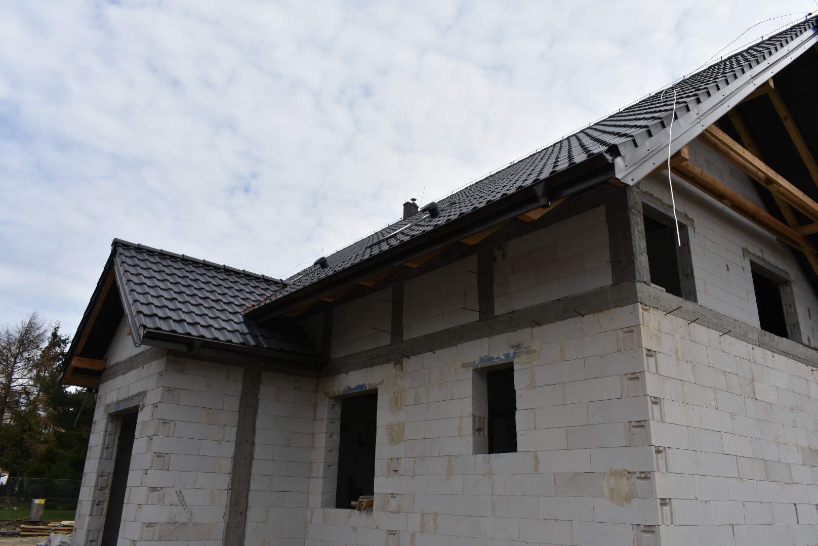 Surowy, dwukondygnacyjny dom z białych bloczków, z ciemną dachówką, w trakcie budowy. Widoczne okna bez szyb, drewniane elementy konstrukcji dachu.