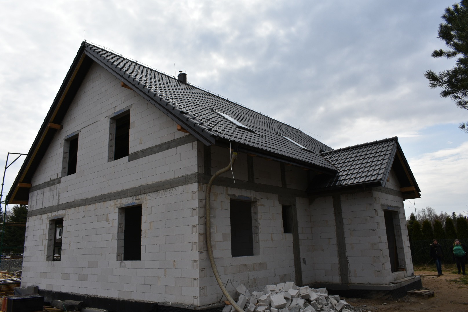 Dwukondygnacyjny dom w stanie surowym otwartym z czarną dachówką, widoczne pustaki, nadproża, fragment elewacji i składowane materiały budowlane.