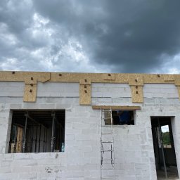 RAF - BUD USŁUGI OGÓLNOBUDOWLANE RAFAŁ MILCZAREK - Wznoszenie ścian domu z betonu komórkowego. Widoczne otwory okienne, drewniane nadproża, drabina i pracownik wewnątrz budynku. Pochmurne niebo w tle.