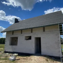 Stan surowy domu z białych pustaków w Lucynowie Dużym. Widoczne okna, drzwi i czarny dach z papy. Na ziemi leżą materiały budowlane, a obok budynku stoi drabina.