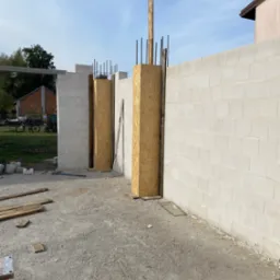 Budowa ściany z bloczków betonowych z drewnianymi szalunkami i wystającymi prętami zbrojeniowymi. Widoczne narzędzia i materiały budowlane na placu budowy.