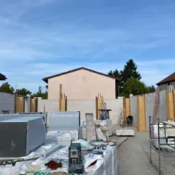 Budowa domu w toku: wzniesione ściany z betonu komórkowego, widoczne materiały izolacyjne Premium Grafit, narzędzia i pracownik w tle. Jasne niebo.