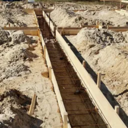 Przygotowany wykop pod fundamenty z drewnianym szalunkiem i zbrojeniem. W tle widoczny dom jednorodzinny i zieleń. Kadr z góry.