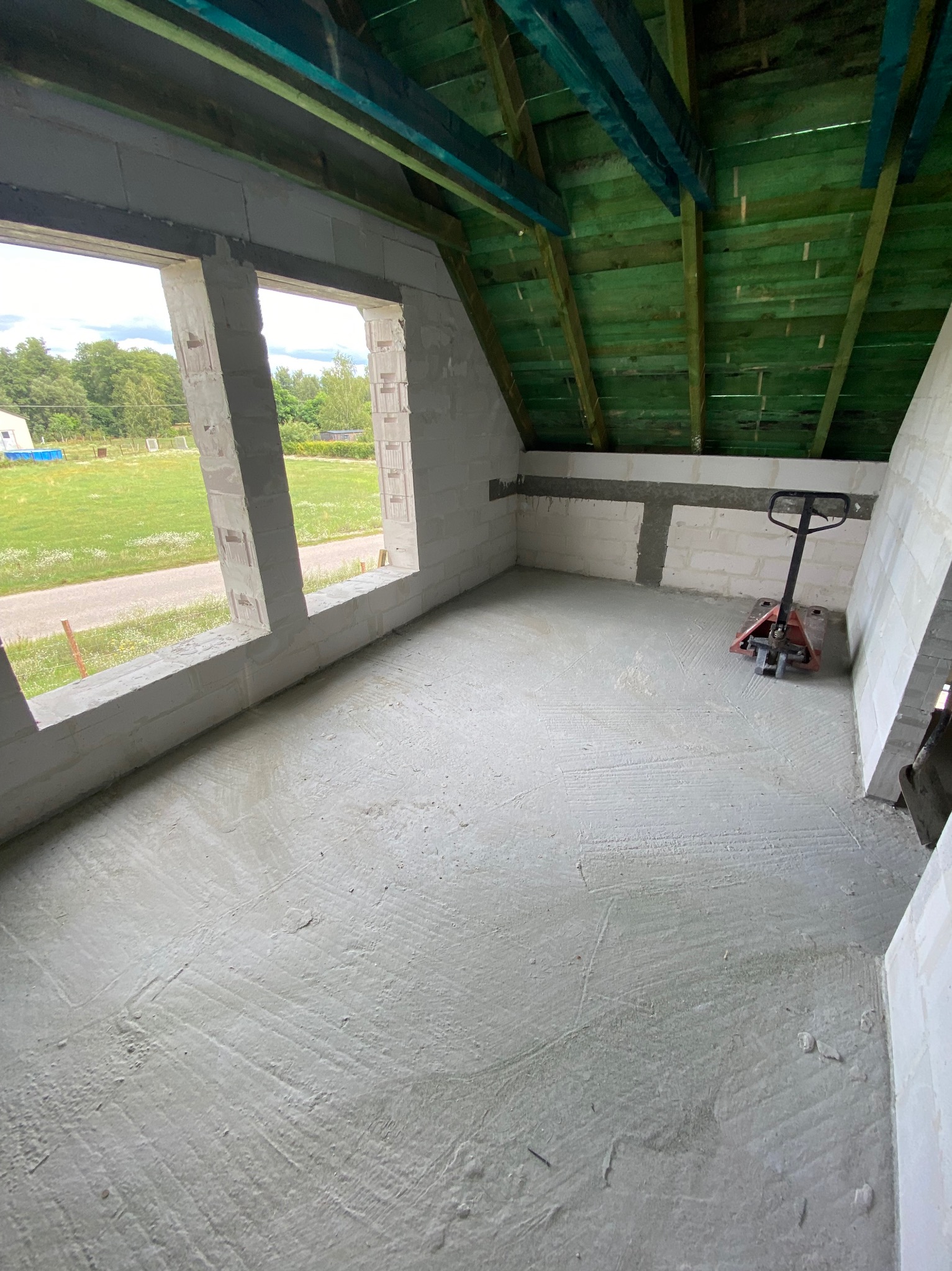 Surowe wnętrze poddasza z oknami, świeżo wylana posadzka, ściany z białych bloczków, zielona drewniana konstrukcja dachu. W tle widok na zewnątrz.