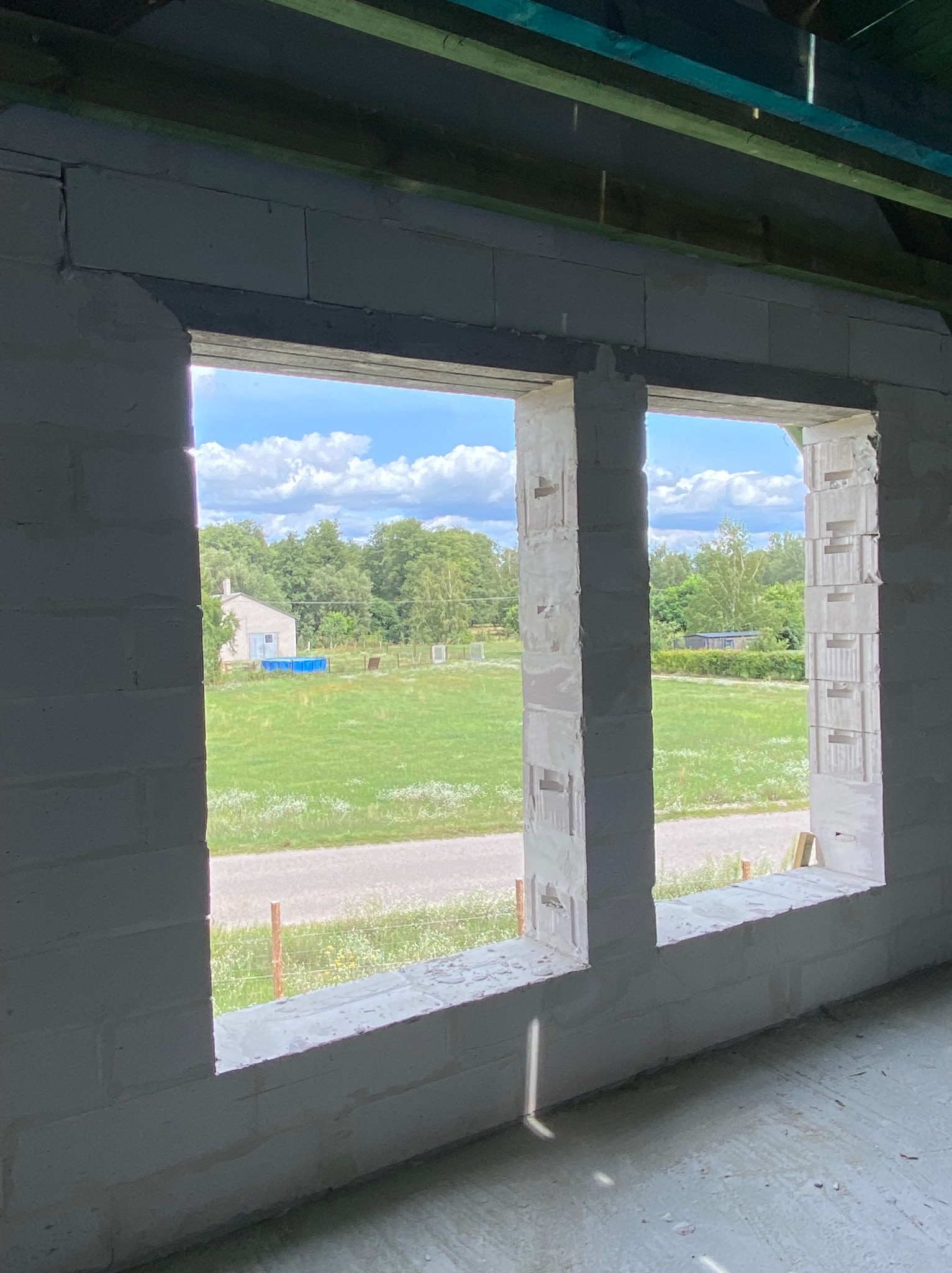 Surowy mur z betonu komórkowego, widok przez dwa otwory okienne na zielony krajobraz z domem i błękitnym basenem. Budowa w toku, surowe wykończenie.