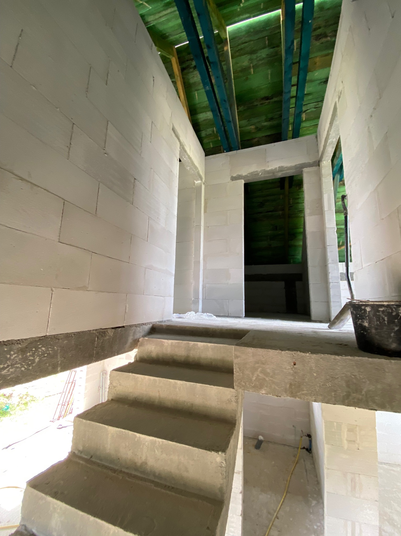 Wnętrze domu w trakcie budowy: białe ściany z betonu komórkowego, betonowe schody, drewniana konstrukcja dachu z widocznymi belkami, wiadro i narzędzia budowlane.