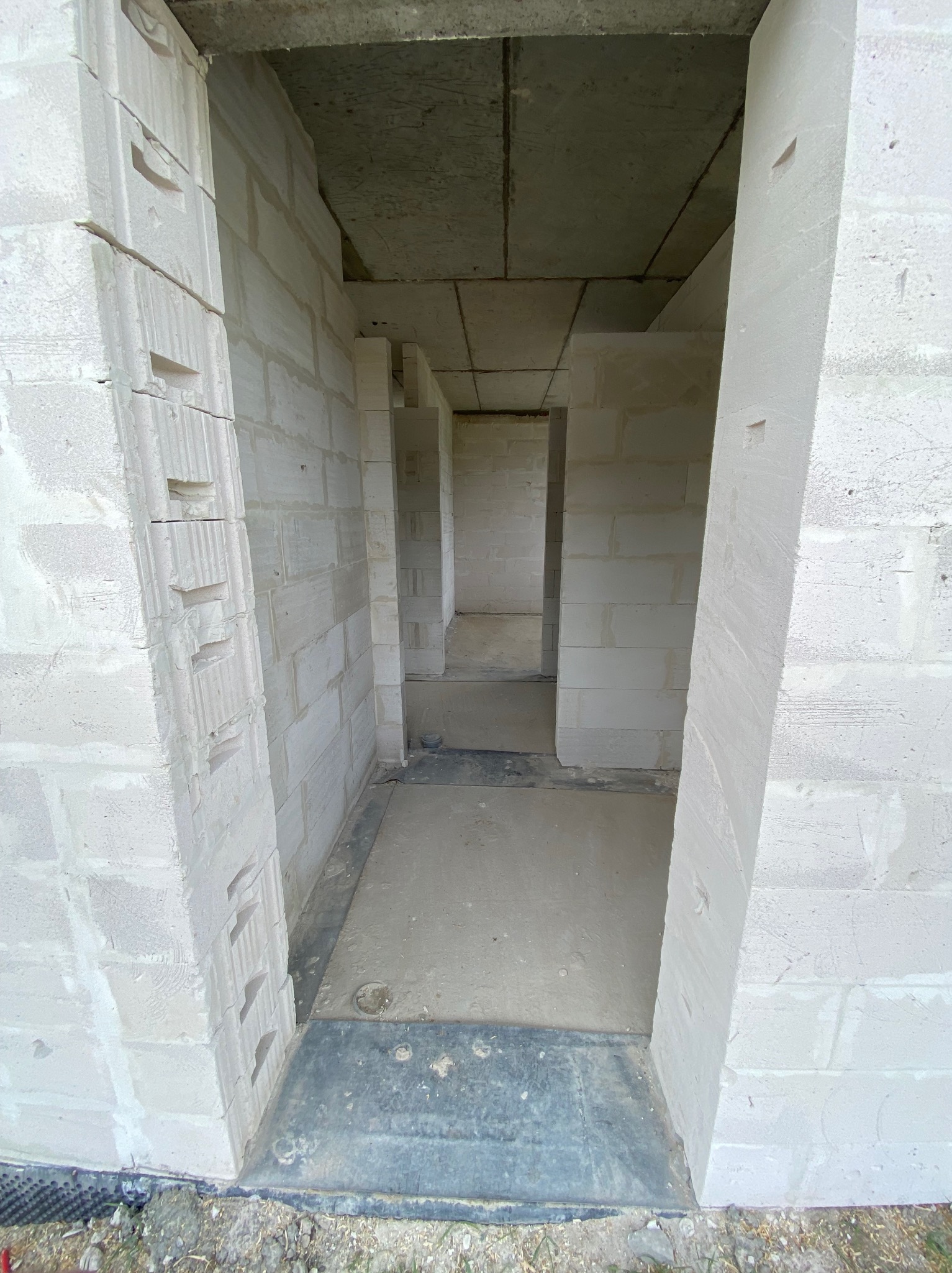 Wnętrze surowego domu z białych bloków, widok z poziomu podłogi na kolejne pomieszczenia. W tle widoczne surowe betonowe stropy.