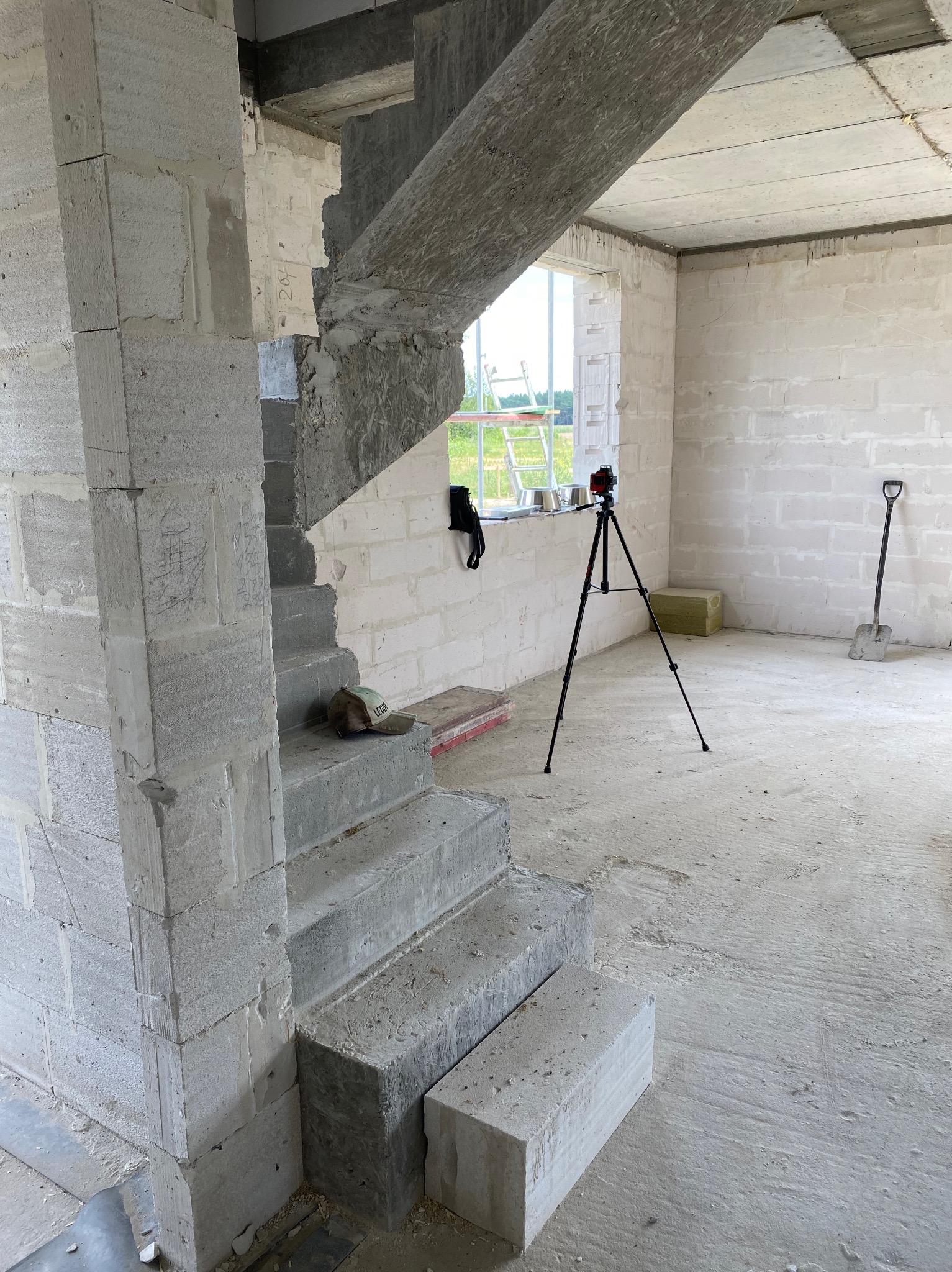 Surowe schody betonowe w trakcie budowy domu z bloczków, widoczny laser na statywie, łopata i okno z zielonym krajobrazem w tle.