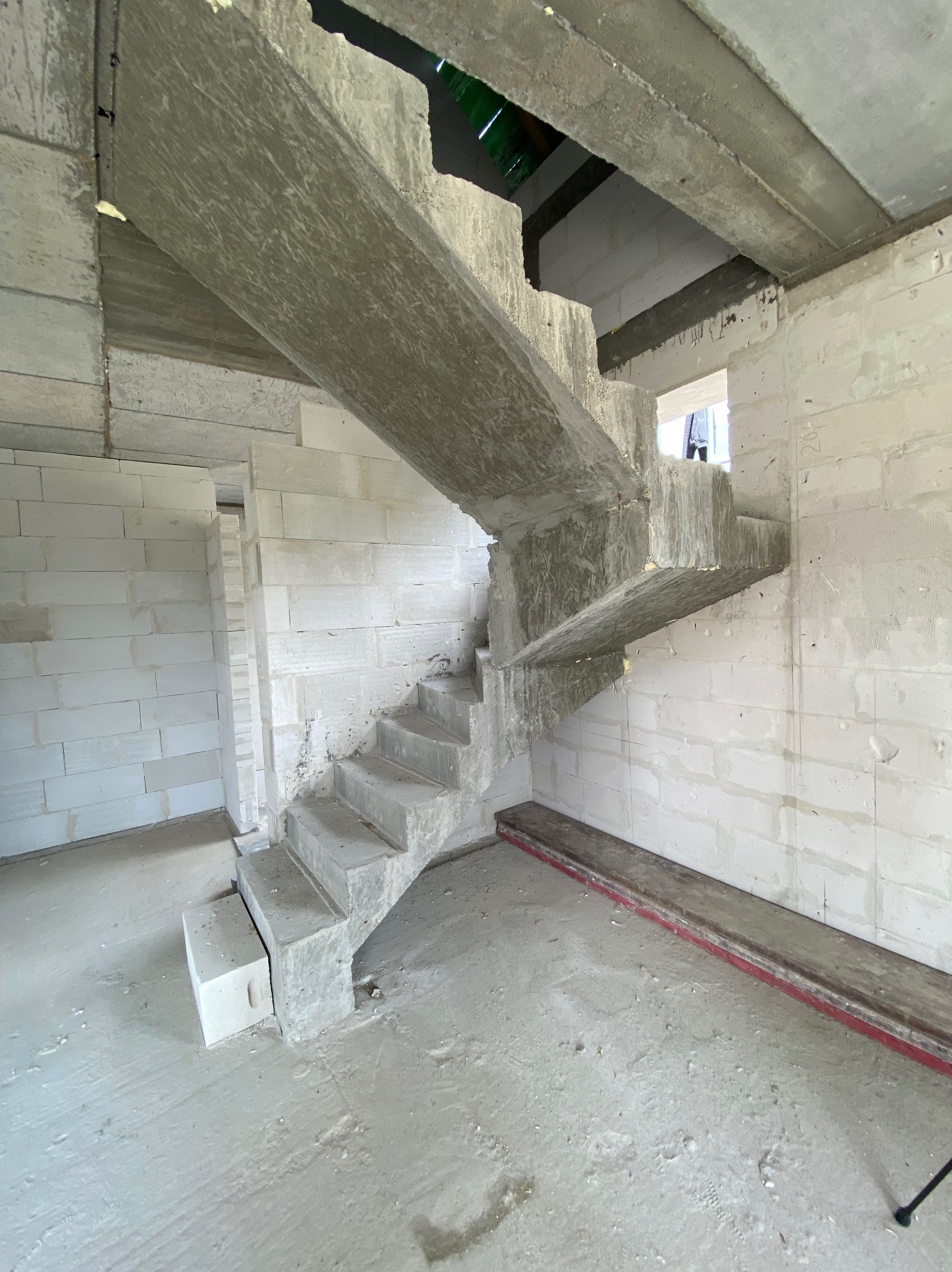 Surowe, betonowe schody w surowym wnętrzu budynku z białych bloczków. Widoczny betonowy strop i fragment okna. W tle widoczne wejście do innego pomieszczenia.